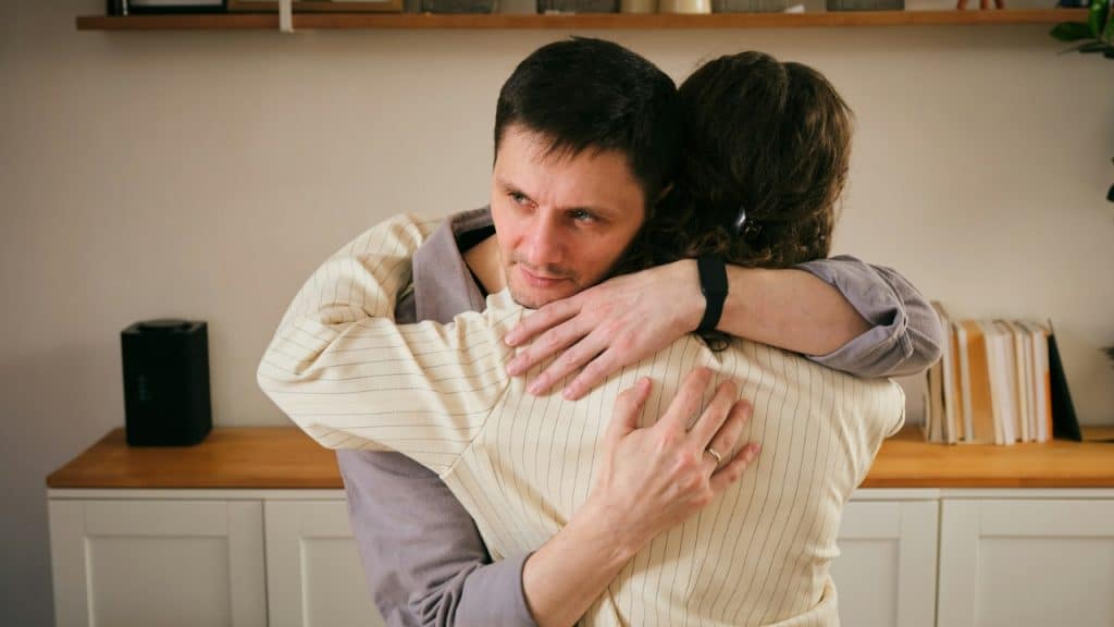 A man with a neutral expression hugs a woman from behind in a brightly lit room.