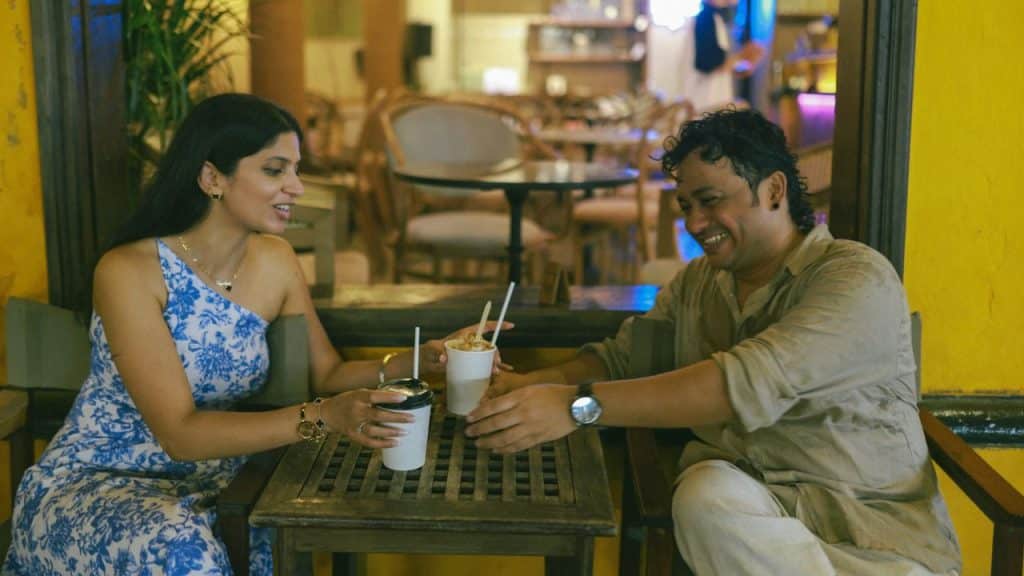 A man and woman sit at an outdoor table laughing and reaching for white cups.