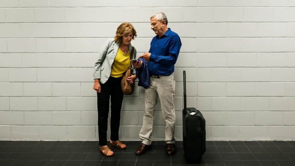 An older man and woman stand by a suitcase and look at a mobile phone.