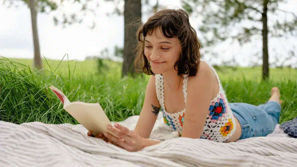 A smiling woman lying on her stomach on a blanket outdoors on grass while reading a book.