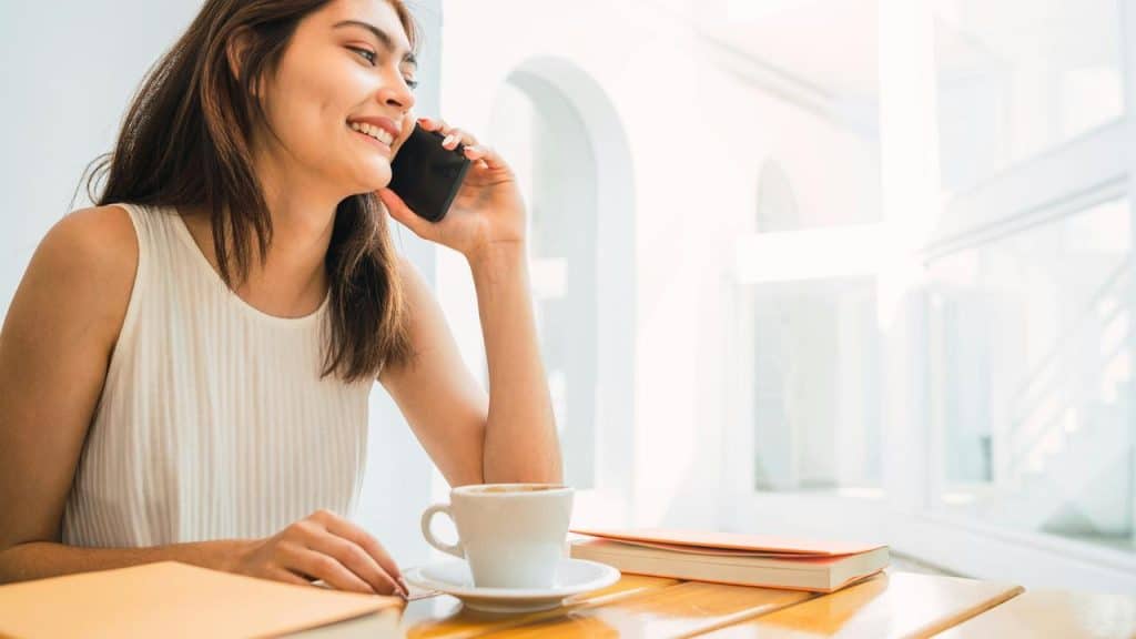 A woman smiling while talking on a phone at a café table with a coffee.