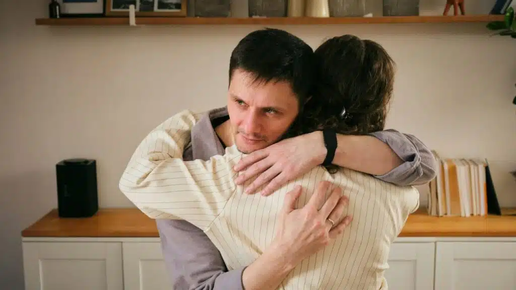 A man with a neutral expression hugs a person whose back is turned to the camera.