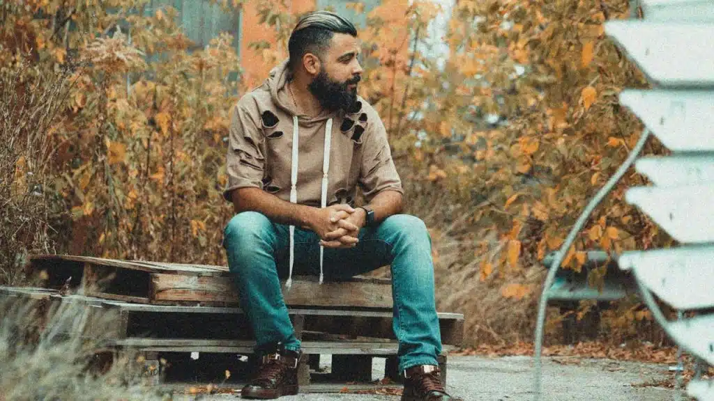 A bearded man sits on wooden pallets outdoors, surrounded by dry brush and autumn leaves.