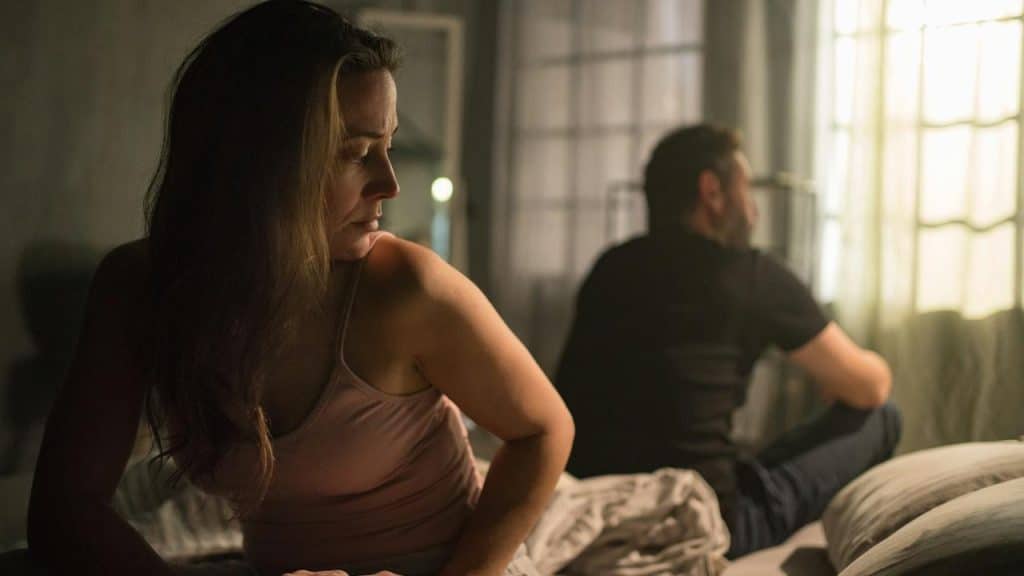 An upset mid-adult woman with her husband in bed behind her—relationship problems.