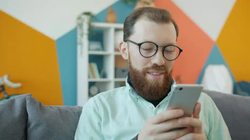 A bearded man wearing glasses smiling while looking down at a smartphone in his hands.