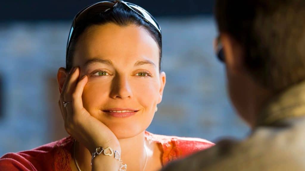 A woman with green eyes rests her chin on her hand while smiling at a person.