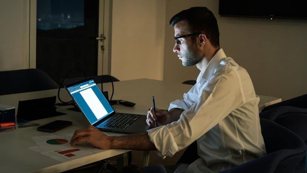 A man working late at night on a laptop with charts on the desk.