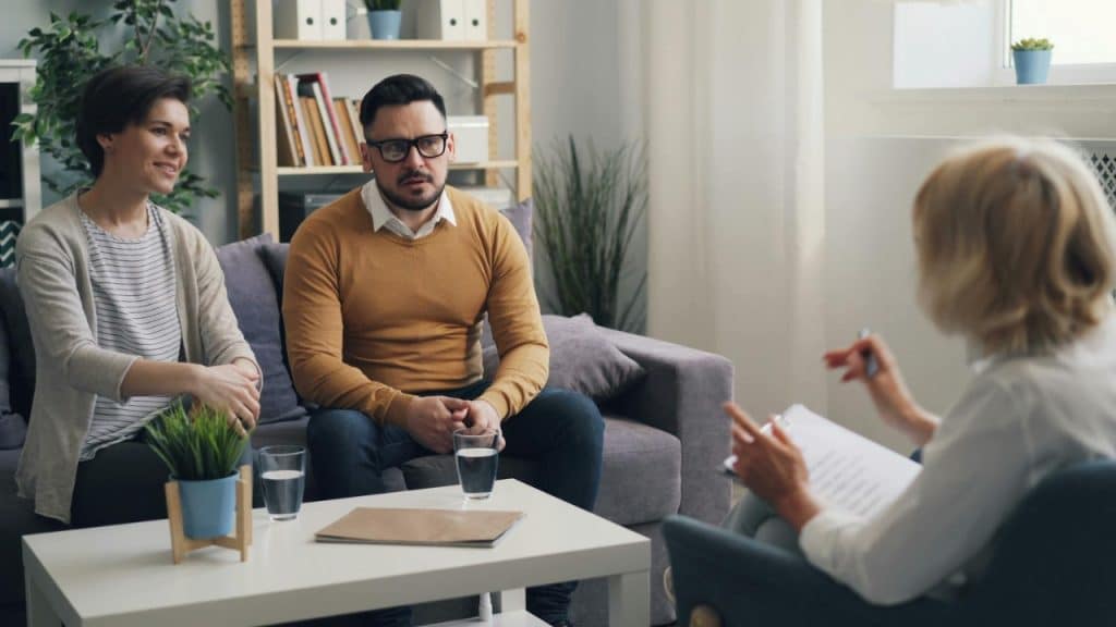 A man and woman sit on a couch facing a person holding a clipboard and pen.Many couples wait until frustration is entrenched before seeking help. By t