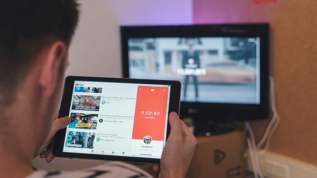 Close-up of hands holding a tablet showing a video app with a television behind.