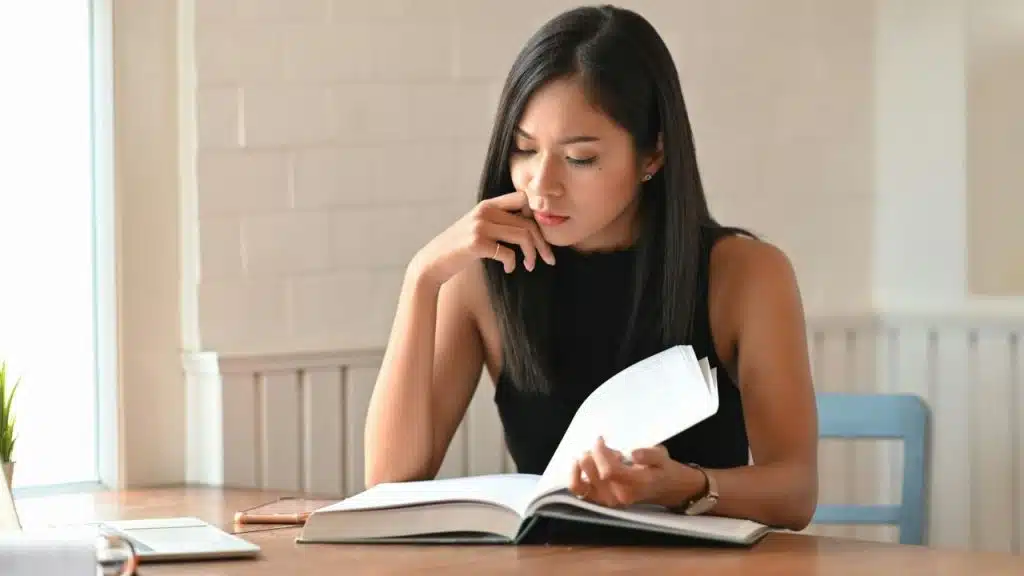 A woman with long black hair sits at a wooden table and reads a large book.
