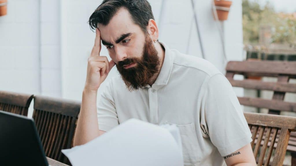 A bearded man sitting outdoors at a wooden table looking intently at a white paper.