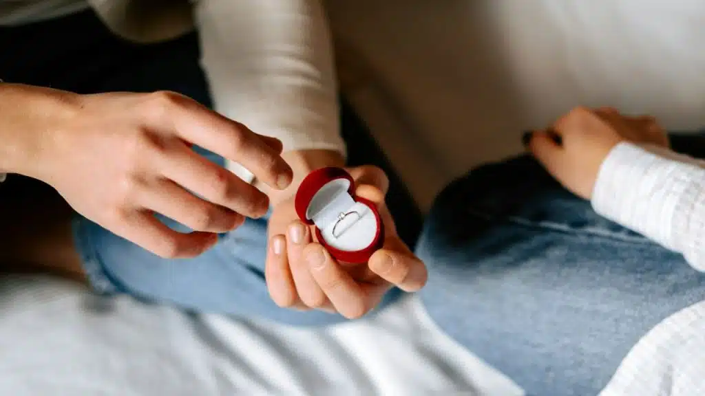 A close-up view of hands holding an open red velvet box containing a small diamond ring.