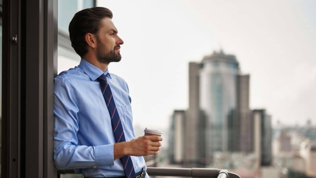 A man in a blue shirt and tie holds a cup while looking over a city.