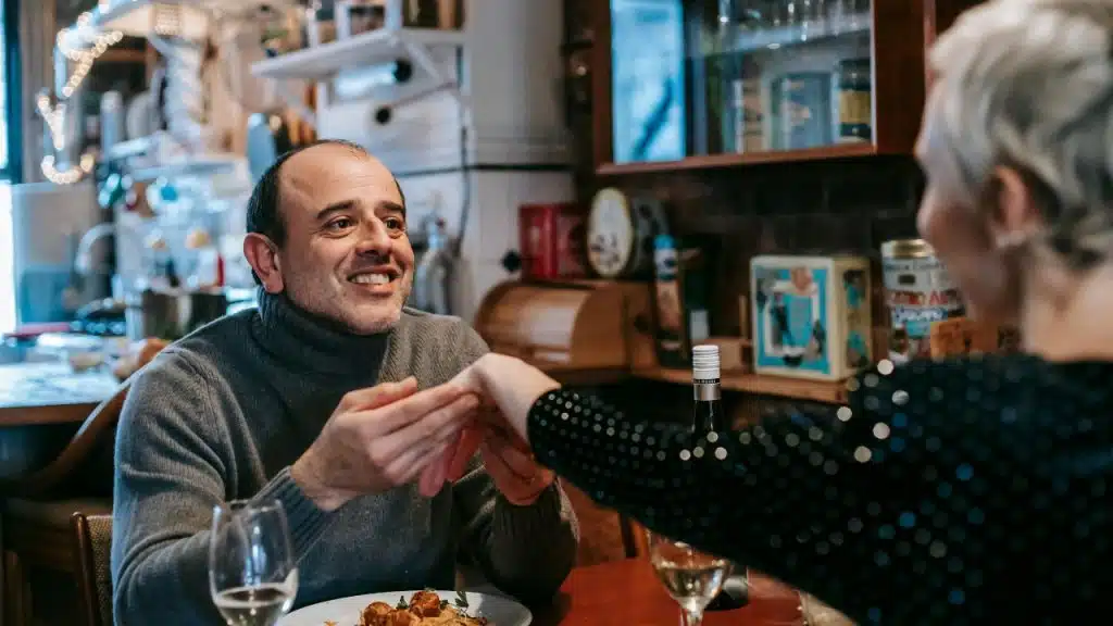 A man in a grey turtleneck holds a woman's hand across a dinner table indoors.