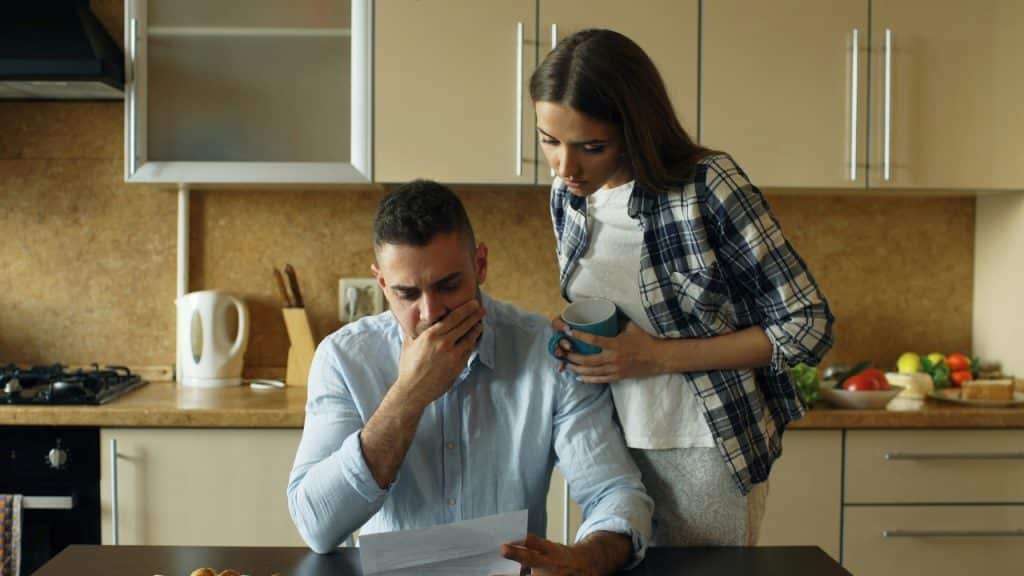 A man sits at a table reading a document while a woman stands behind him.