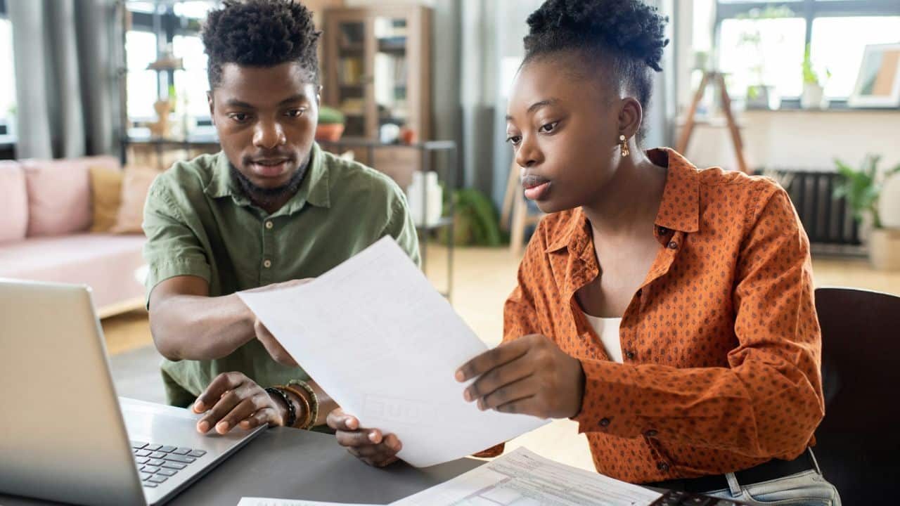 A man and woman sit at a desk together looking at a laptop and paper document.