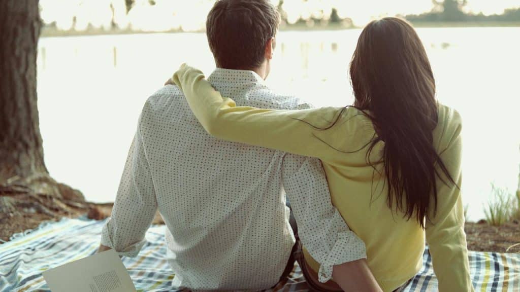 A couple sitting on a blanket by the water with the woman’s arm around the man.