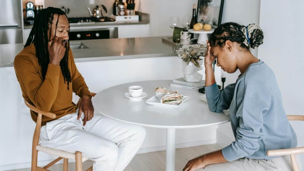 A man and woman sit at a table; the woman rests her head in her hand looking down.