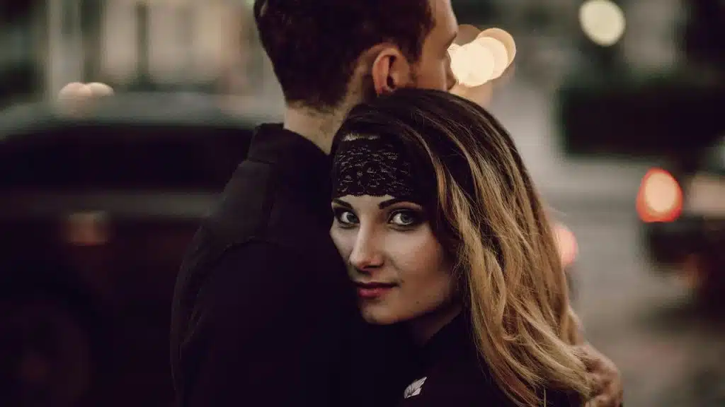 A woman hugging a man on a city street at night, looking toward the camera.