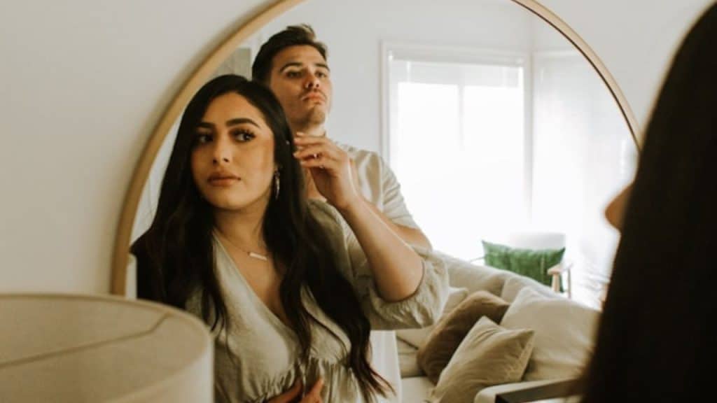 A man stands behind a woman and touches her hair while looking into a mirror.