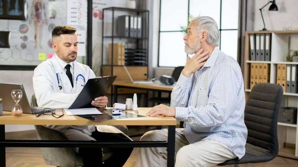 A male doctor in a white coat talks to an older patient at a desk.