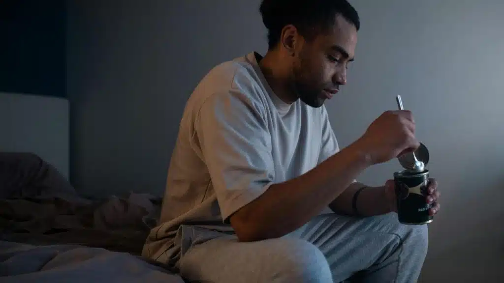 A person sitting on a bed eating from a container with a spoon.