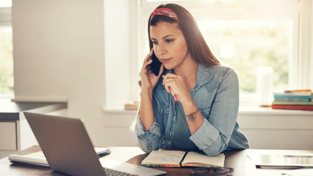 A woman with a red headband talks on a cellphone while looking at a laptop computer.
