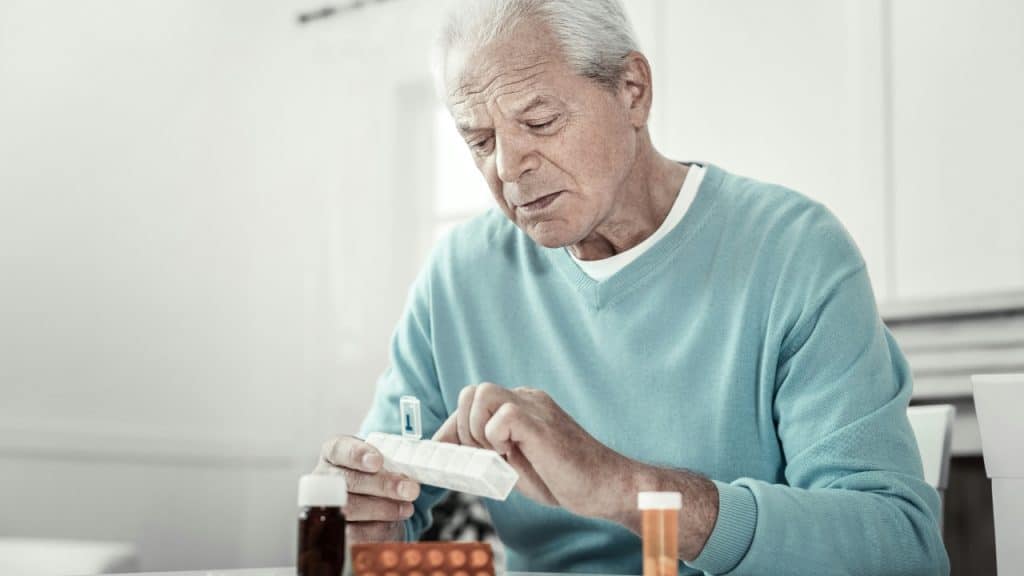 An older man in a blue sweater looks down while organizing pills in a white container.