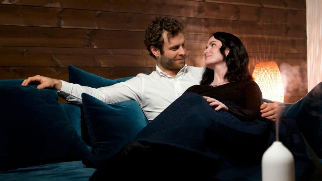 A man and woman sit on a teal sofa, looking at each other while reclining.