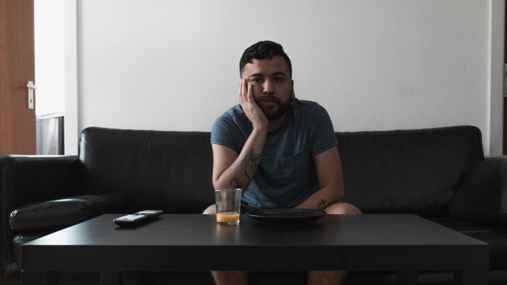 A man with a tattooed arm sits on a sofa behind a dark wooden coffee table.