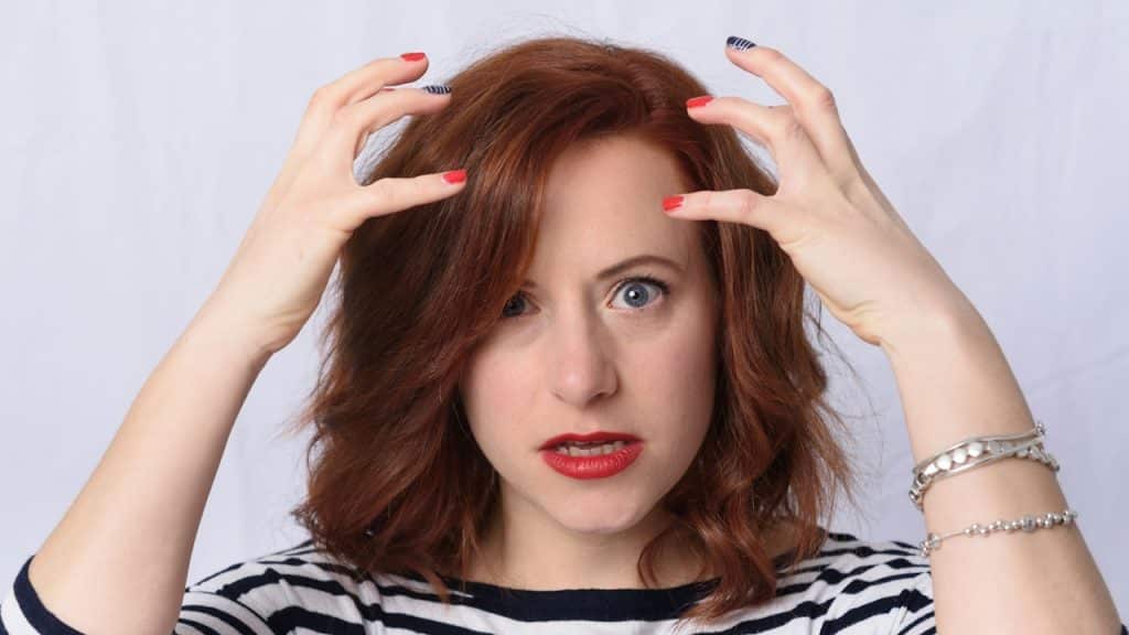 A woman with short red hair looks directly at the camera with a frustrated expression, raising both hands toward her head.
