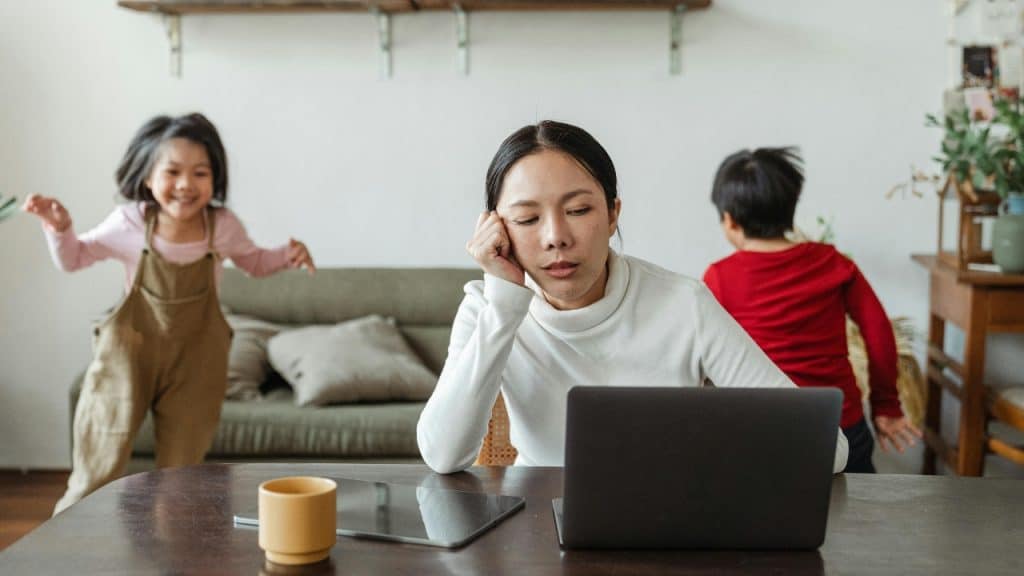 A woman sits at a laptop with a tired expression while two children play behind.