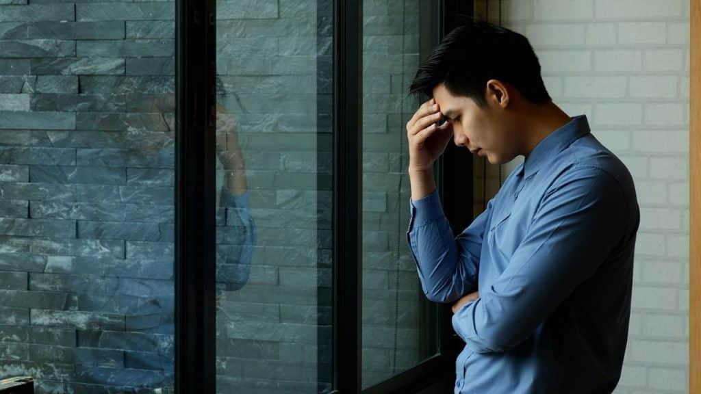 A stressed man in a blue shirt is standing by a window, holding his head in his hand.