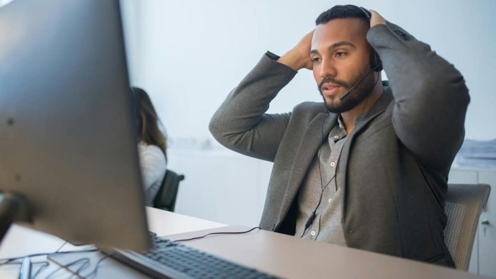 A man wearing a headset sitting at a desk with his hands on his head.