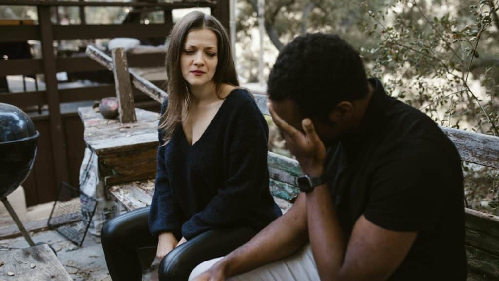 A woman with visible tears sits on a bench next to a man covering his face.