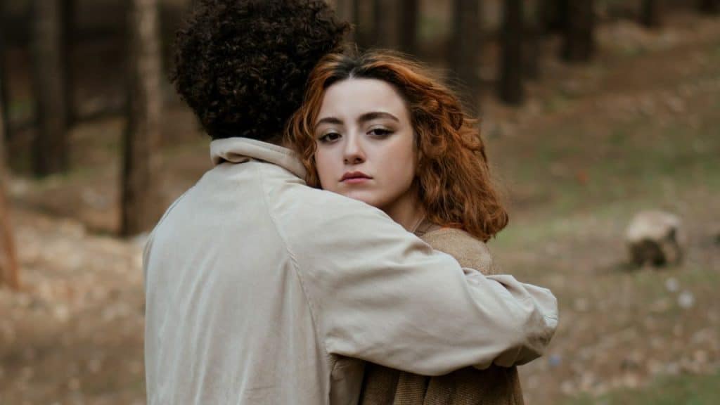 A person with reddish-brown hair embraces another person with dark curly hair in a wooded area