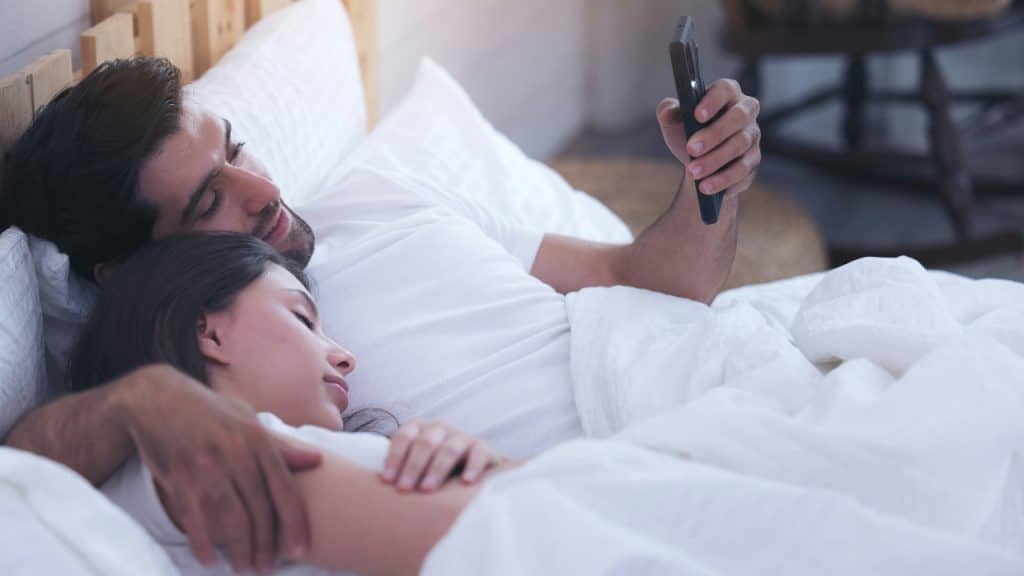 A man lies in bed using a phone while a woman sleeps beside him under linens.