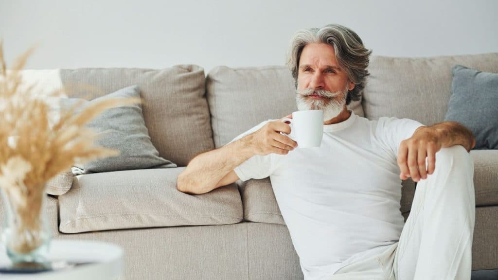 A man with a grey beard sits on the floor by a couch, holding a mug.