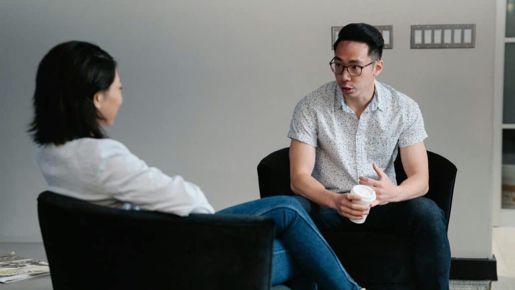 A man in glasses talks while sitting in a chair across from another person.