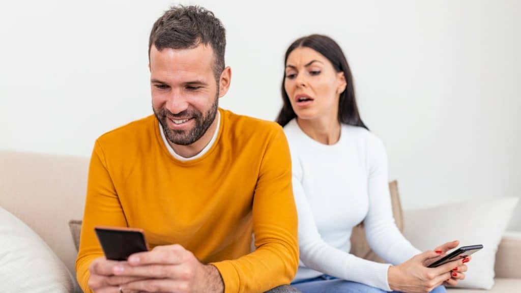 A smiling man looks at his phone while a woman behind him looks on with concern.