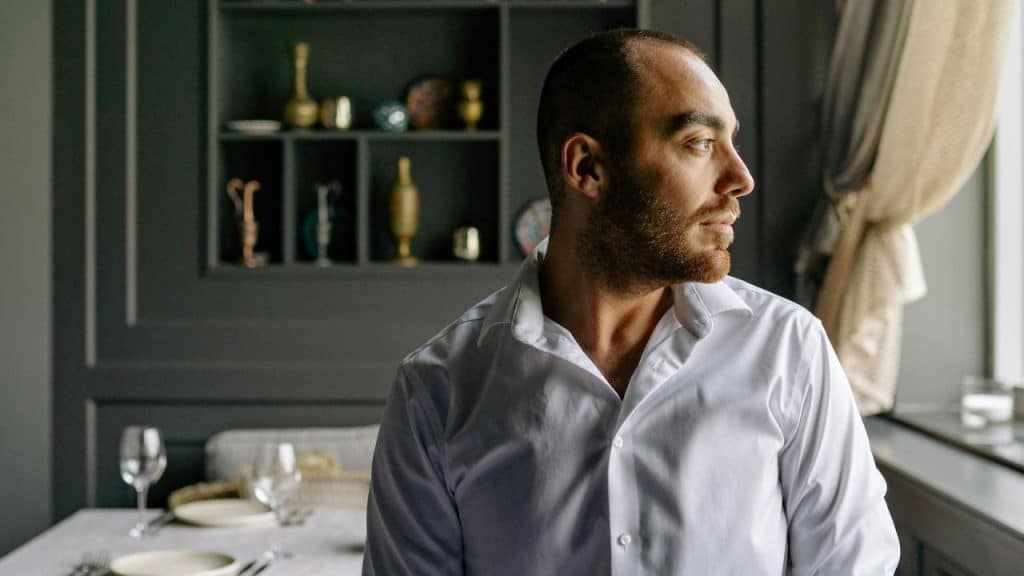 A man in a white shirt looking out a window indoors next to a dinner table.