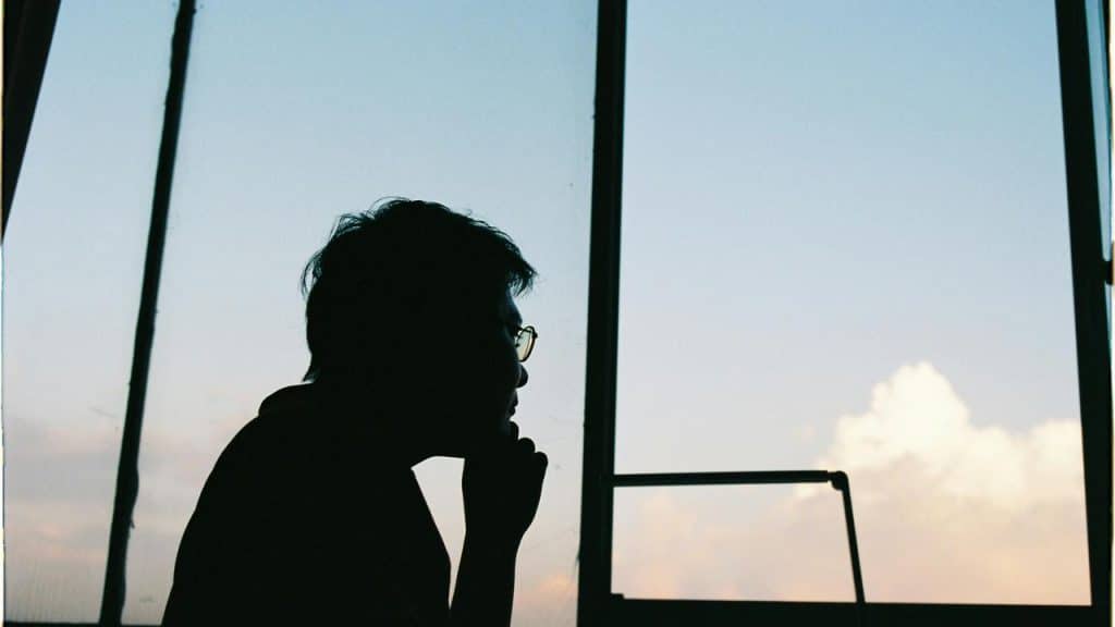 A silhouetted person gazes thoughtfully out a window at the sky.