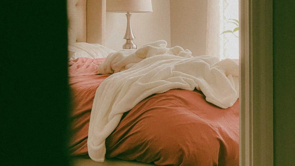 An unmade bed with rumpled white blankets in a softly lit bedroom.