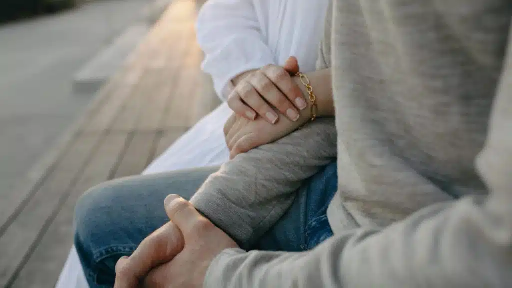 A person’s hand with a gold bracelet rests on another person's arm on a bench.