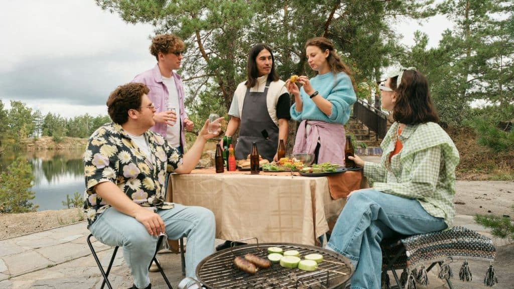 Five people stand and sit around a table outdoors near a lake during a meal.
