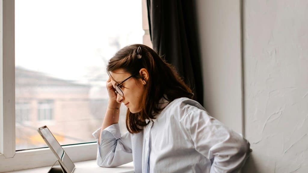 A woman wearing glasses, leaning by a window while looking at a tablet.
