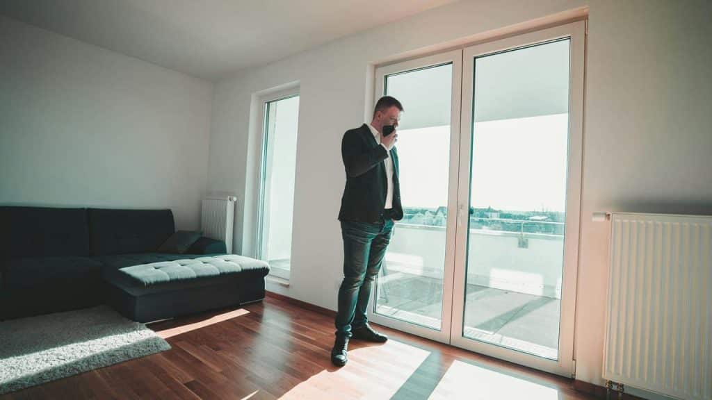 A man stands by a large window in a sparsely furnished room while speaking on his phone.