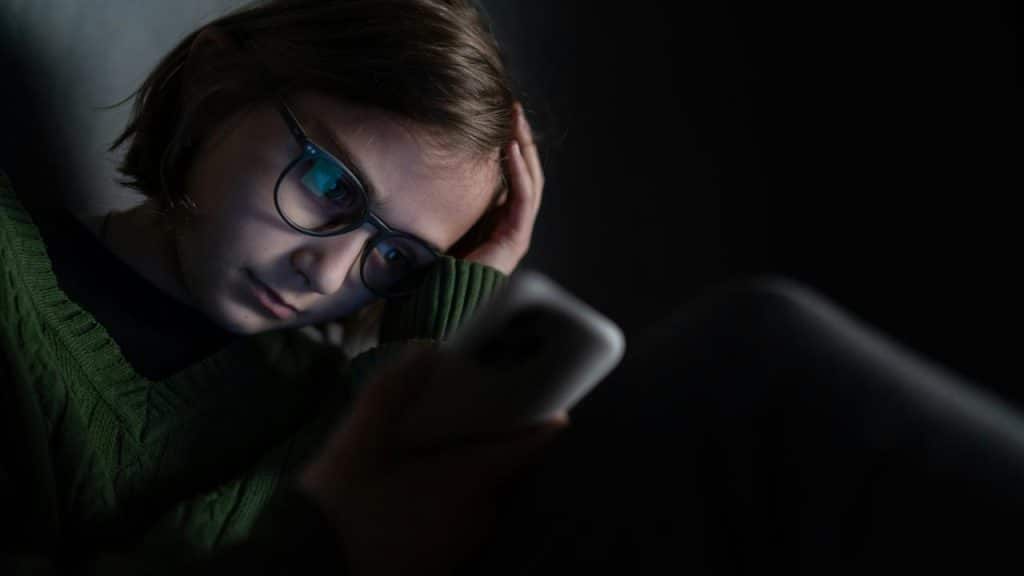 A person wearing glasses, resting their head while looking at a phone in the dark.