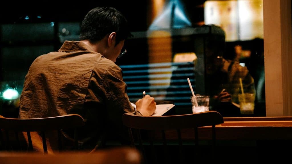 A person in a brown shirt sits at a dark wooden table writing in a notebook.