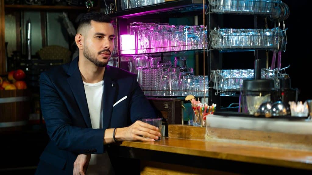 A man in a navy blazer sits at a bar holding a glass near glass shelves.